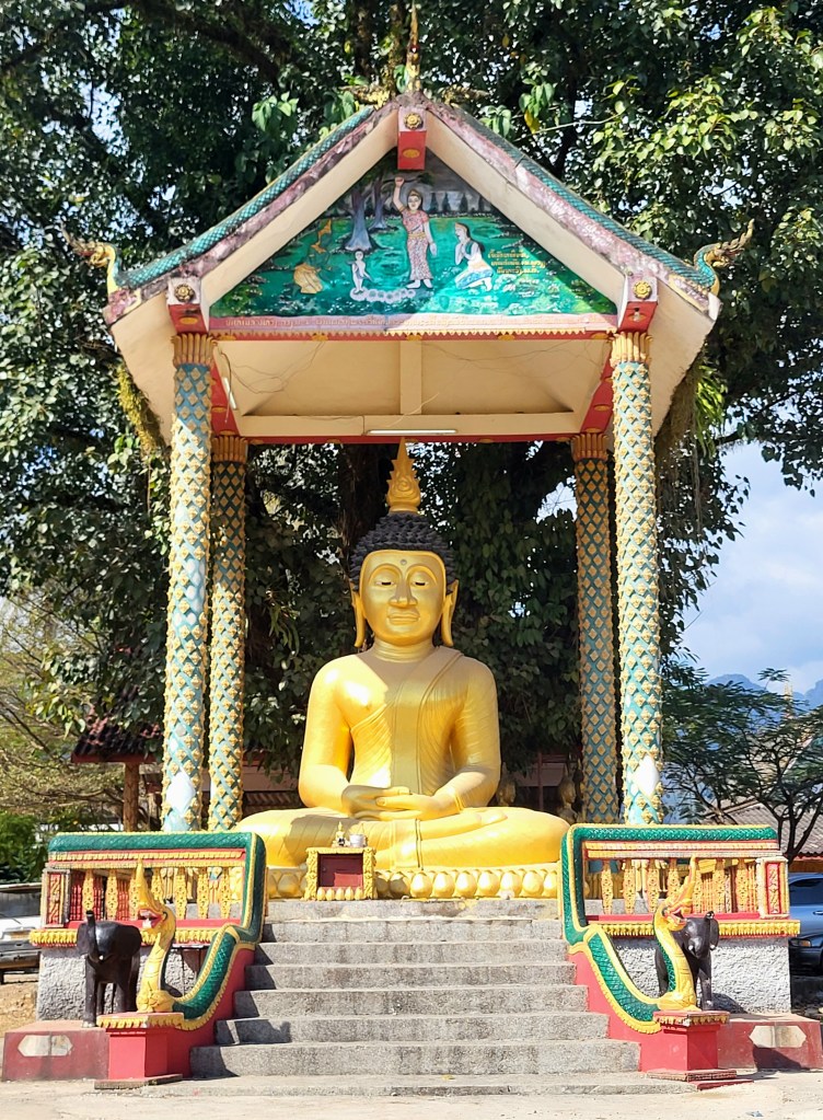 Statue en or d'un Bouddha assis sous un abri décoré, entourée d'éléments artistiques et de la végétation.