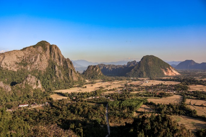 Vue panoramique des montagnes et de la vallée avec des champs et un chemin sinueux sous un ciel bleu.