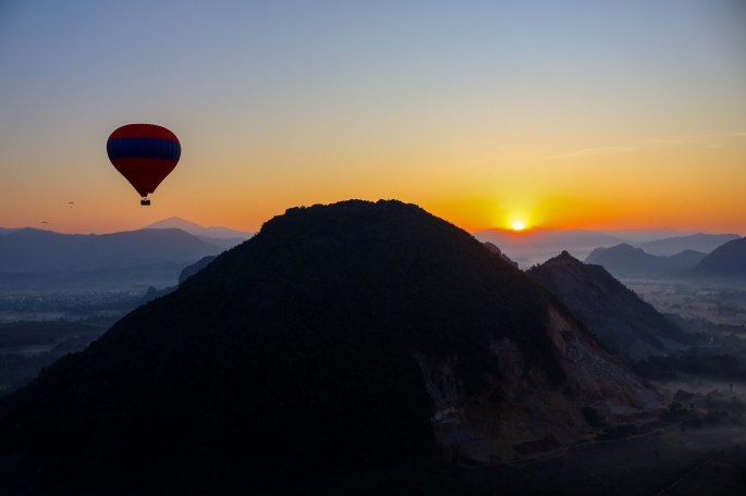 Montgolfière survolant une colline au lever du soleil, avec des montagnes à l'arrière-plan.