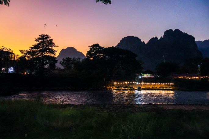 Coucher de soleil au bord d'une rivière avec des montagnes en arrière-plan et des lumières scintillantes sur la rive.